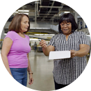 Job Area_161x161_Distribution Center Two women are standing in a brightly lit industrial space. The woman on the left is wearing a pink shirt and jeans, while the woman on the right is in a striped shirt and dark pants, holding a clipboard and pointing at it. They appear to be engaged in conversation.