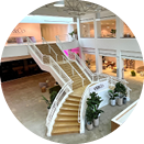 A modern interior space featuring a grand staircase with wooden steps and white railings. The open layout includes multiple levels with glass partitions, potted plants, and well-lit areas, creating a bright and inviting atmosphere.