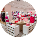 A circular image showing the interior of a lingerie store. Mannequins display colorful lingerie sets on a round table, surrounded by neatly organized drawers and shelves filled with more lingerie items. The store has a bright, modern decor with pink accents.